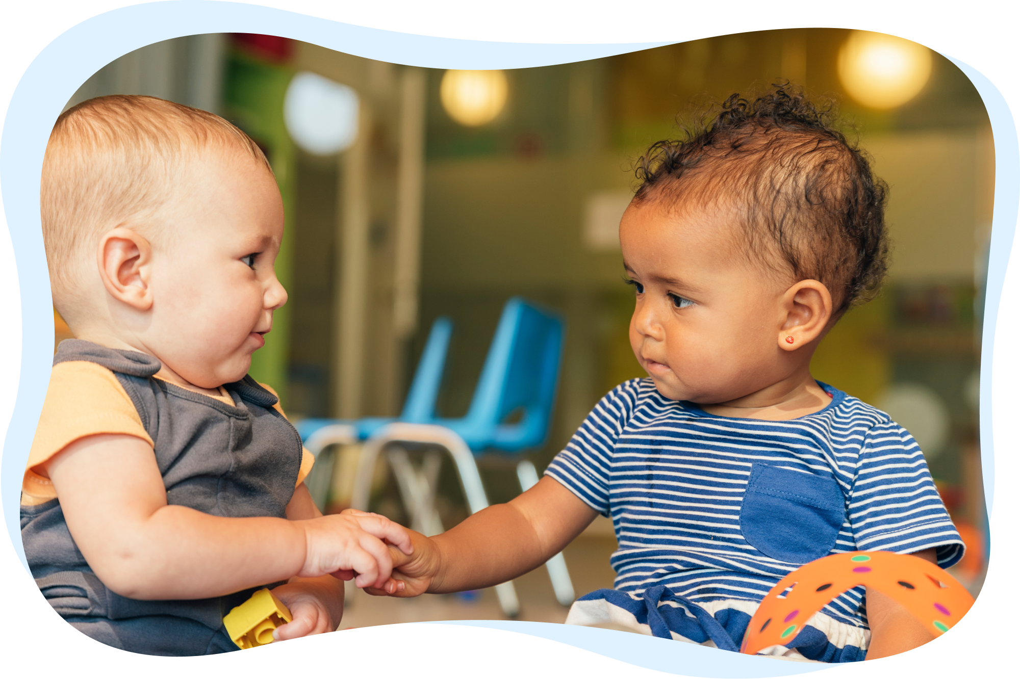 Two babies sitting and looking at each other while holding hands.