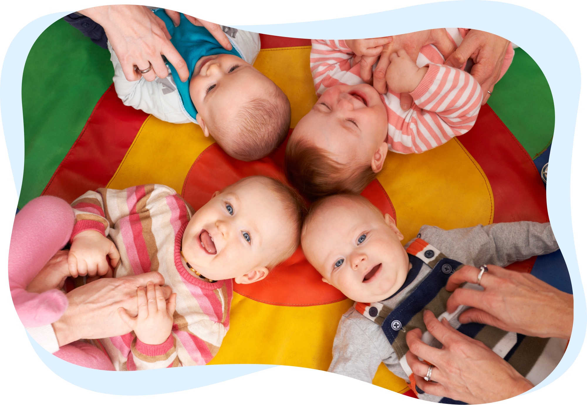 Four babies lying in a circle on a multicolored play mat, smiling and playing.