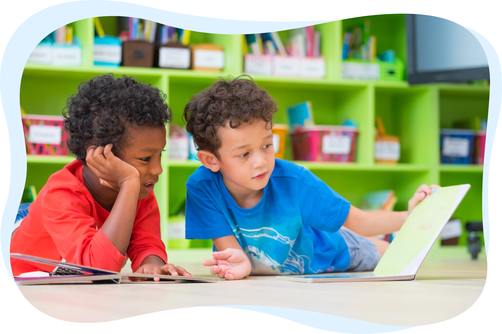 Two young boys lying on the floor, reading picture books together in a classroom.