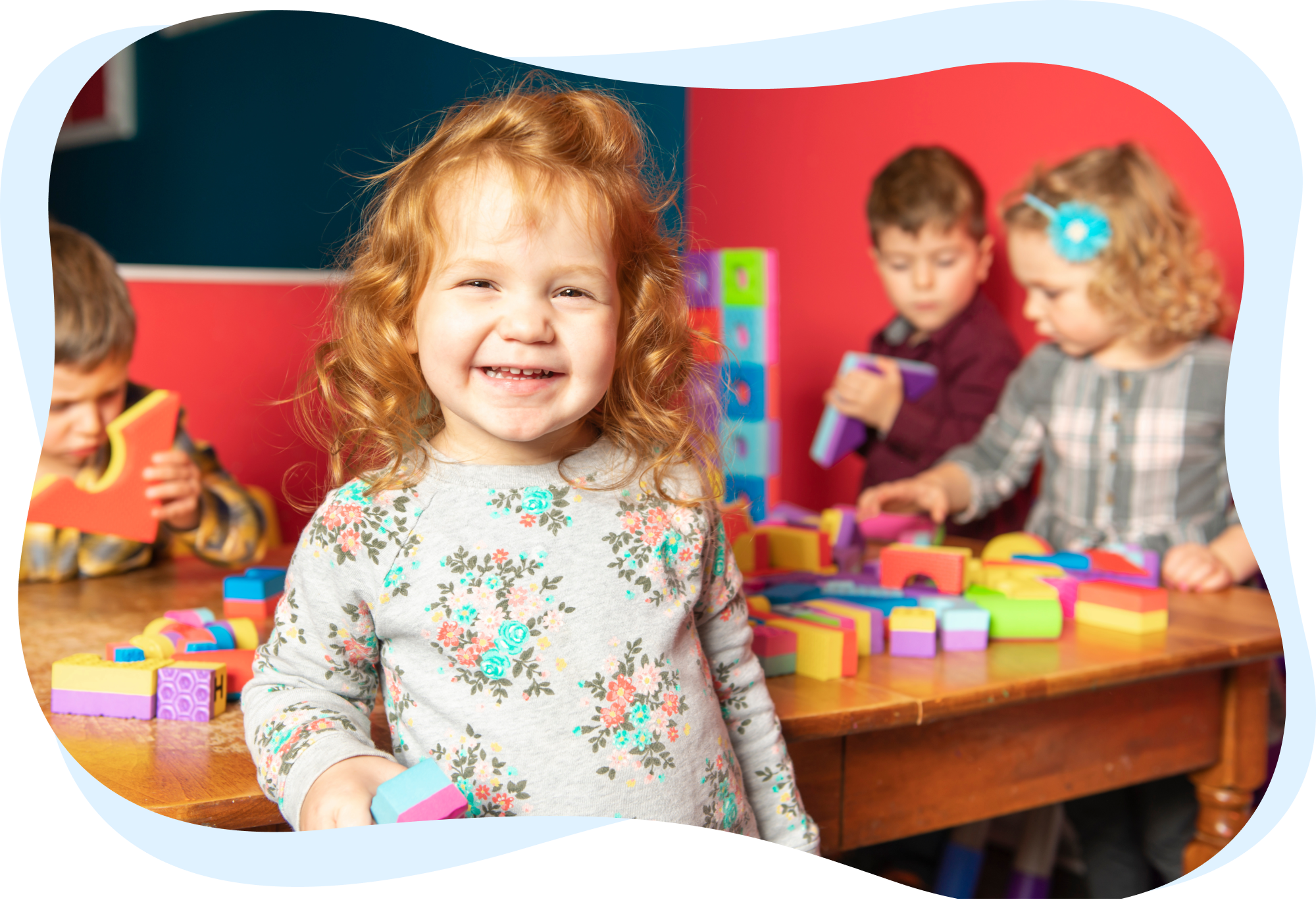 A smiling young girl holding a block, with other children playing with blocks in the background.