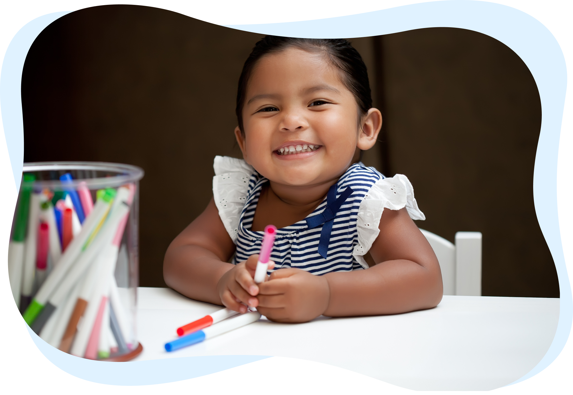 A smiling young girl holding markers, sitting at a table with a container of colorful markers.