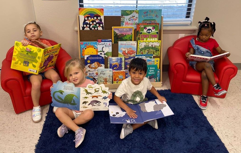 Four Pre-Scholar children sit in a cozy reading area, each holding a book, with a bookshelf full of colorful books behind them.