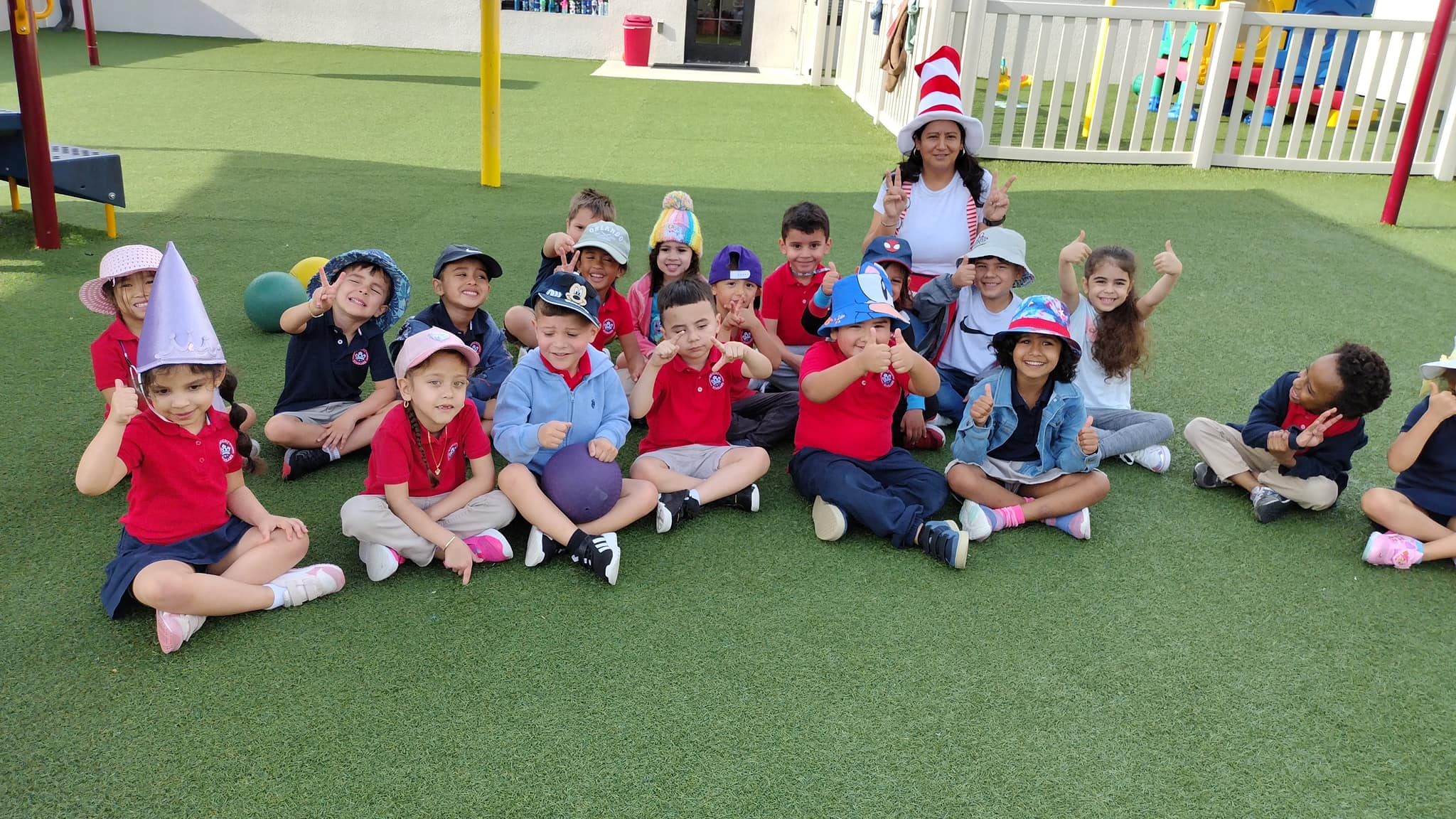 A group of Pre-Scholars at Bright Scholars sit outdoors on grass, wearing various hats, with a teacher in a Cat in the Hat hat.