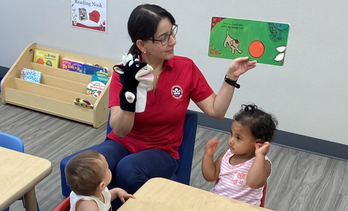 A teacher reads a book to young children at a small table, using a cow puppet to engage them during storytime at Bright Scholars.